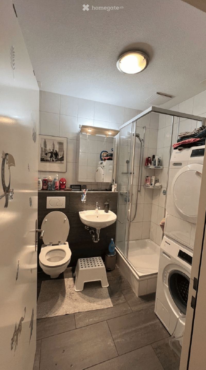 Modern bathroom with a toilet, sink, mirror, shower, and a washing machine. Includes a small stool and a picture frame on the wall.