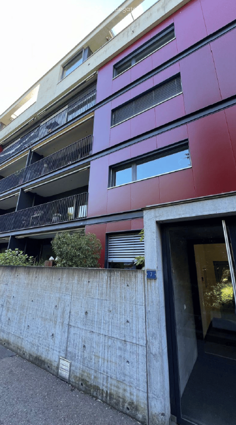 Modern multi-story building with red and gray facade, multiple balconies, and a concrete wall in the foreground.