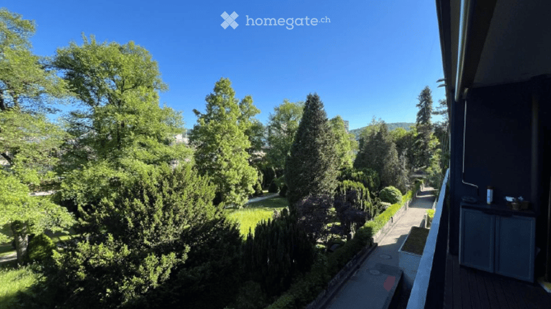 Balcony overlooking a lush, green garden with tall trees and neatly trimmed hedges. The balcony has a wooden deck and a small storage cabinet.
