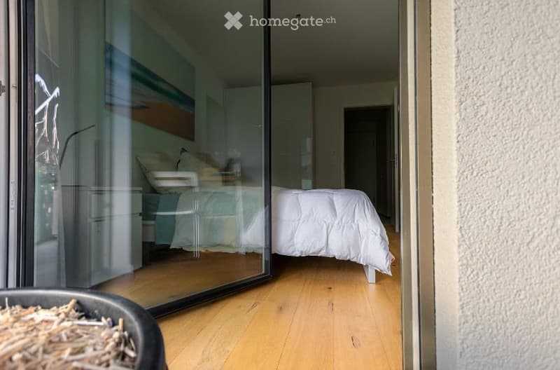 Modern bedroom with a large glass sliding door leading to a balcony, featuring a neatly made bed with white bedding, a nightstand with a lamp, and a small chair near the bed.