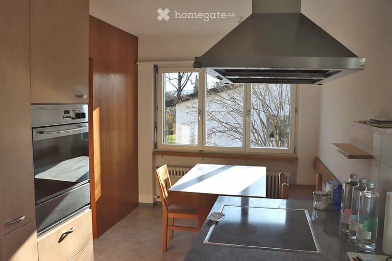Modern kitchen with light wooden cabinets, stainless steel appliances, an induction stove, and a dining area with a table and chair.