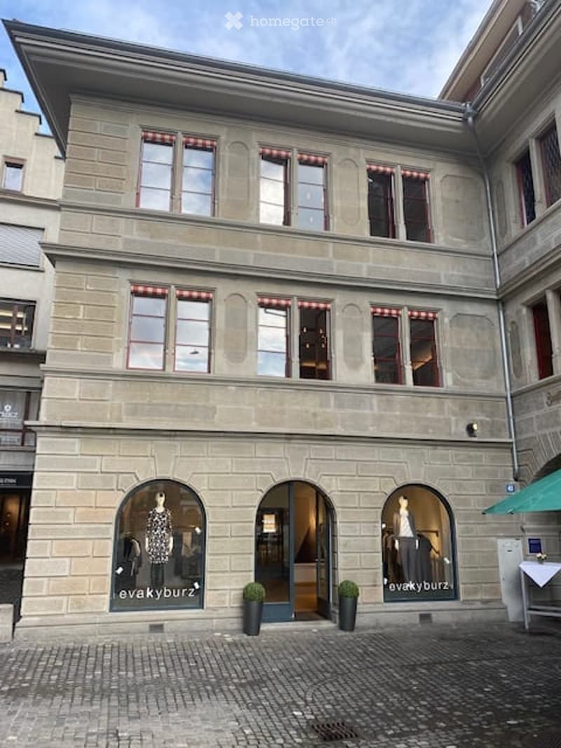 Three-story building with stone facade, large windows with red shutters, and a storefront with mannequins and signage reading "evakyburz".