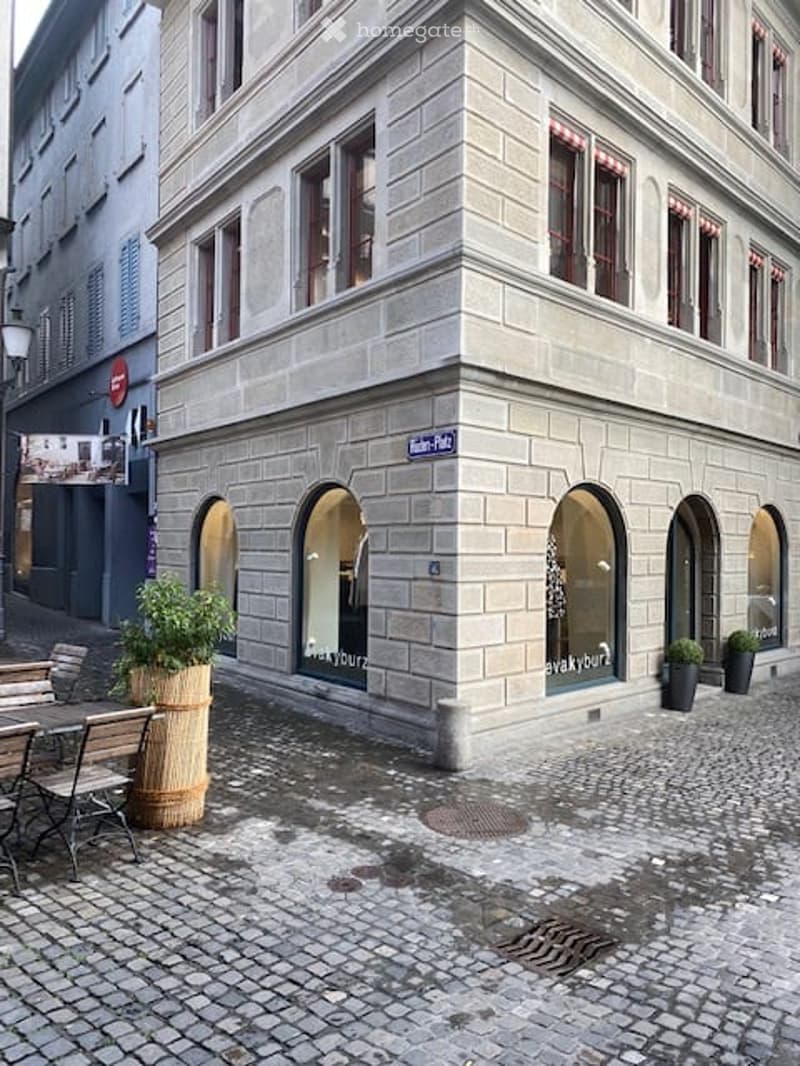Corner building with light gray stone facade, arched windows, and storefronts. Potted plants and outdoor seating are visible on the sidewalk.