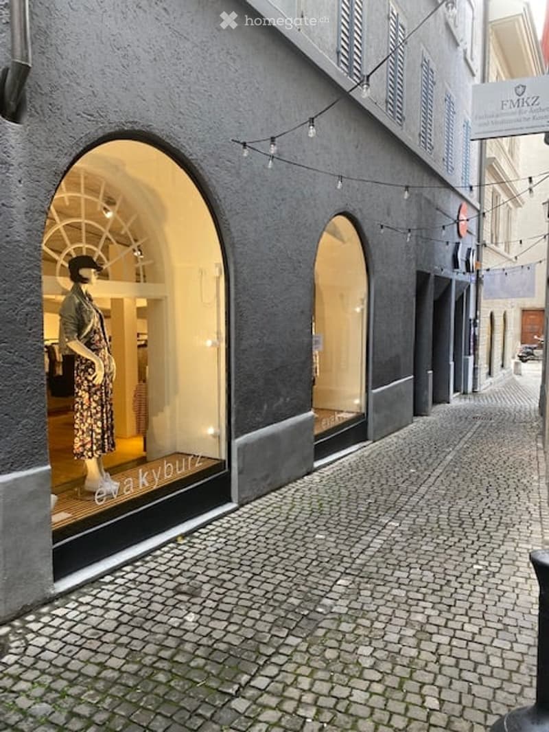 Exterior view of a building with a storefront featuring large arched windows and a mannequin dressed in clothing displayed inside. The building has a textured grey facade and is situated on a cobblestone street.