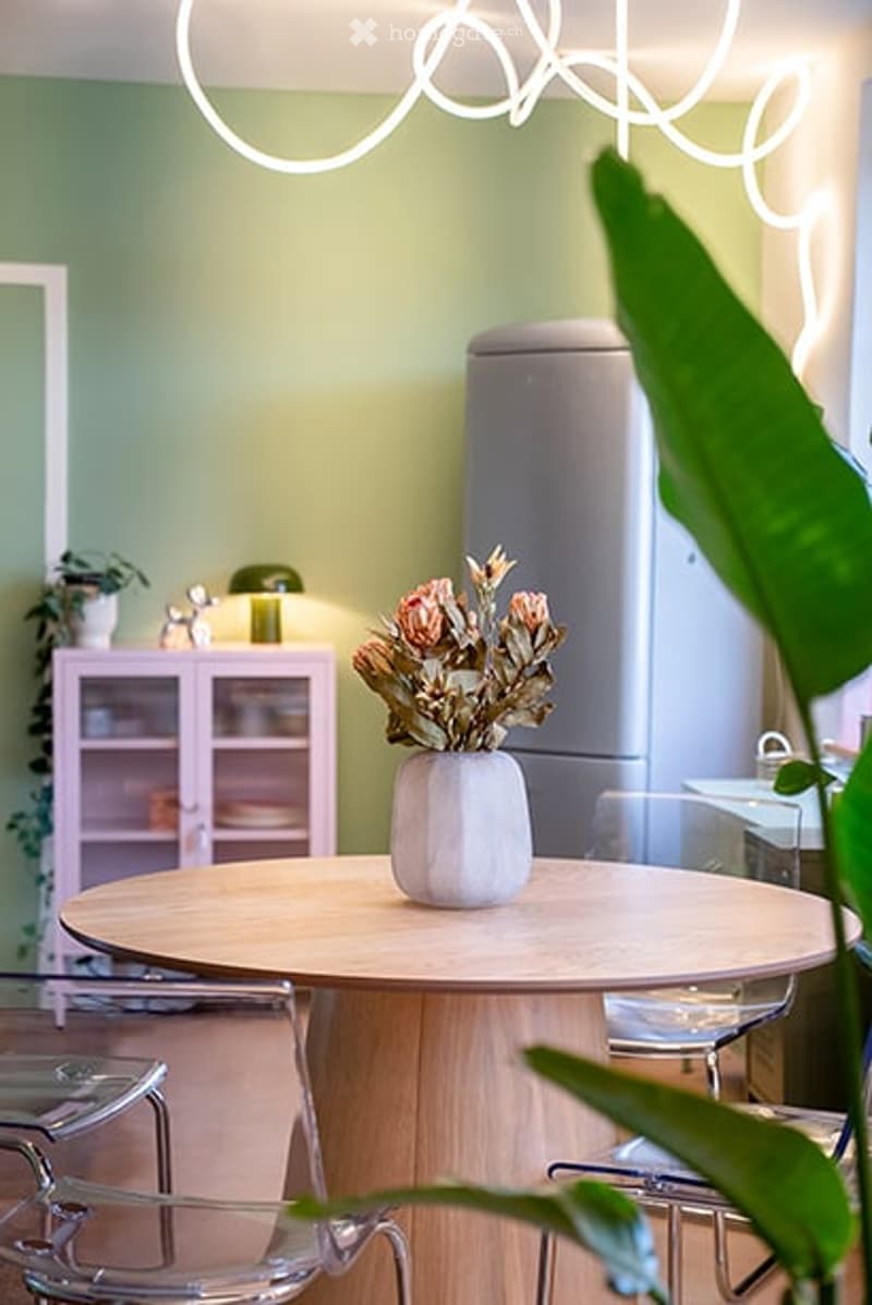 A modern kitchen area with a round wooden table, clear chairs, a white cabinet, and a vase with flowers on the table. A refrigerator is visible in the background, along with a potted plant and decorative lighting.