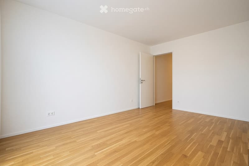 Empty room with light wooden flooring and white walls, featuring an open doorway leading to another room.