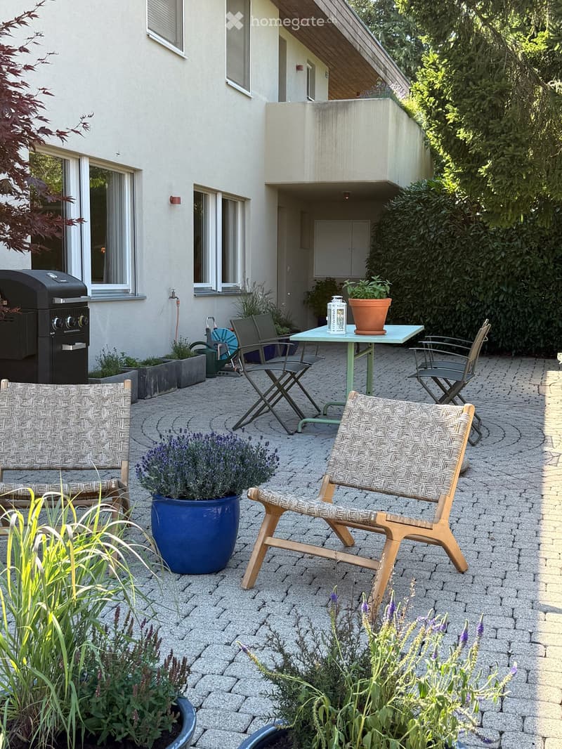 A well-maintained patio area with a stone-paved floor, featuring a small table with four chairs, potted plants, and a grill on the left side.
