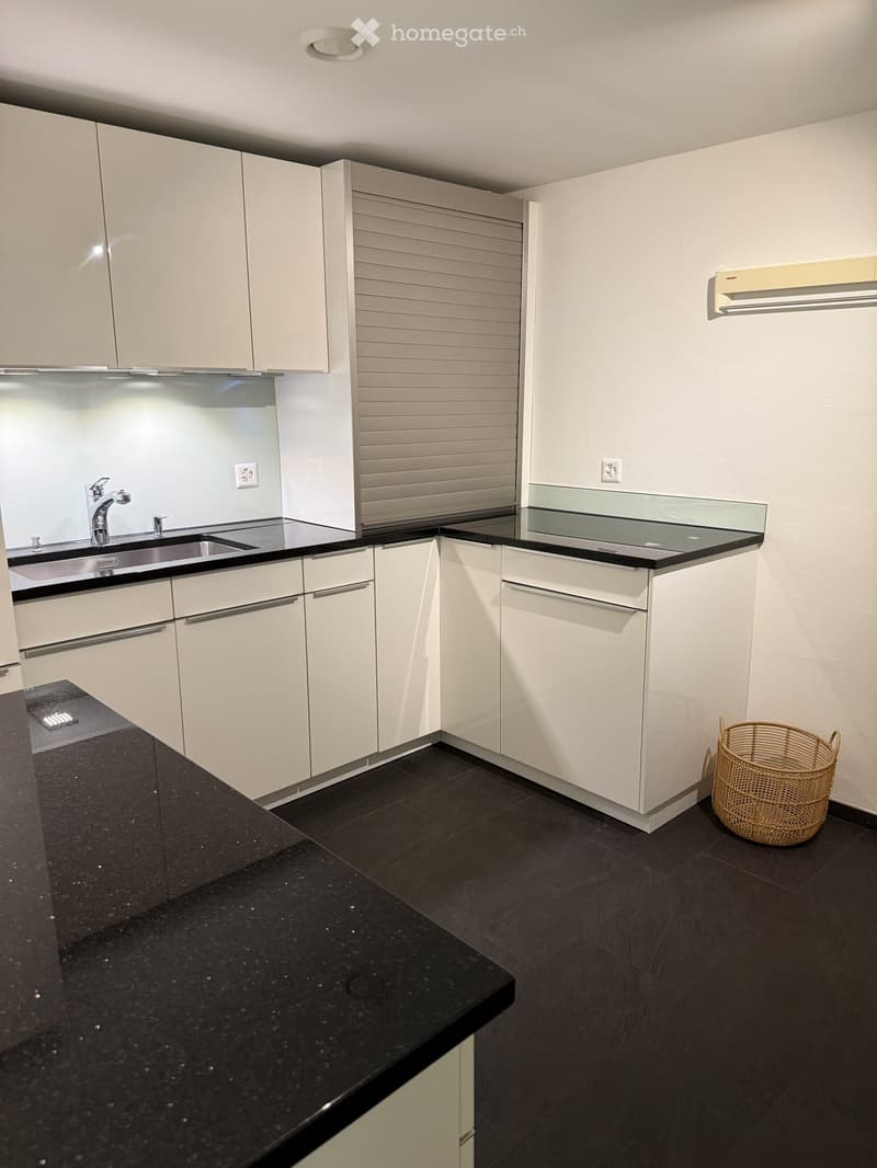 Modern kitchen with white cabinets, black countertops, and a black tiled floor. Features a sink with a modern faucet, a wicker basket on the floor, and a sleek design.