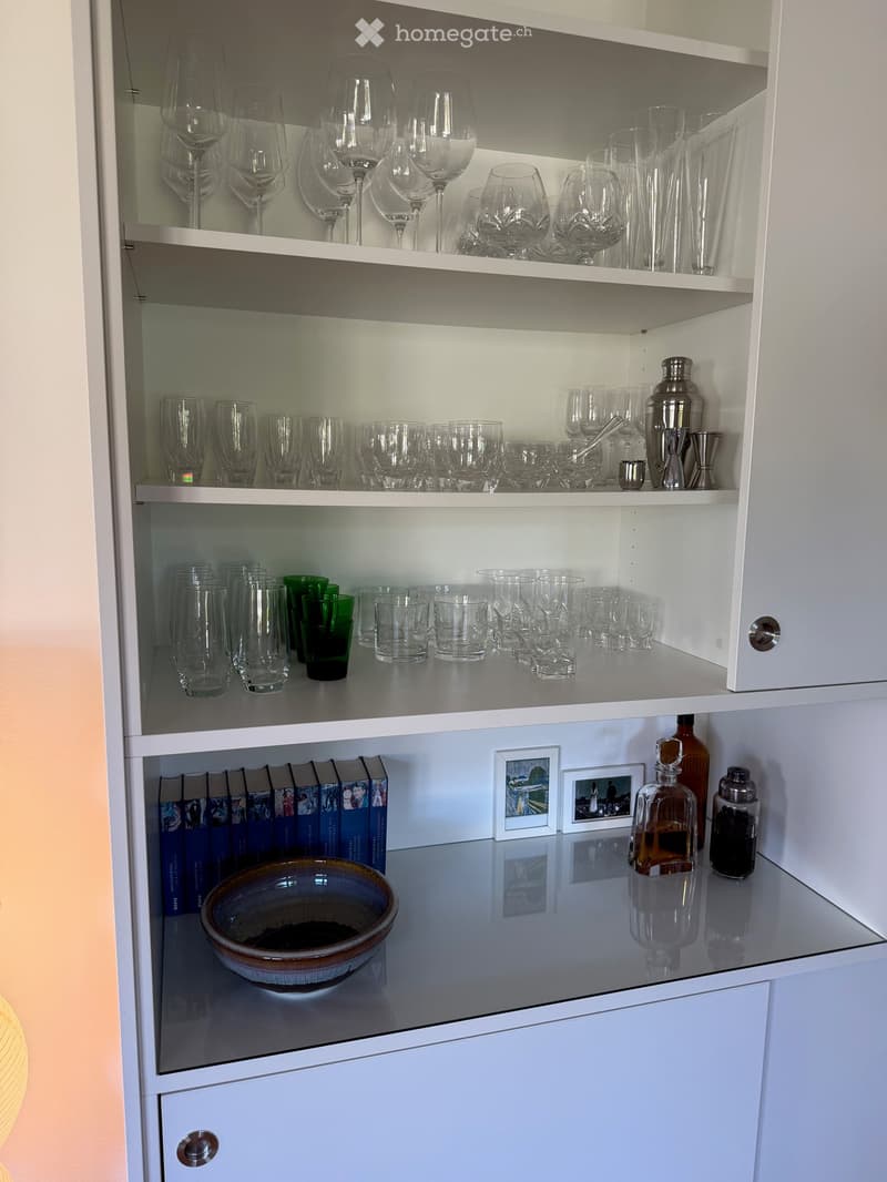 White kitchen cabinet with multiple shelves filled with wine glasses, cocktail glasses, shot glasses, and a few bottles. A bowl and books are placed on the lower shelf.