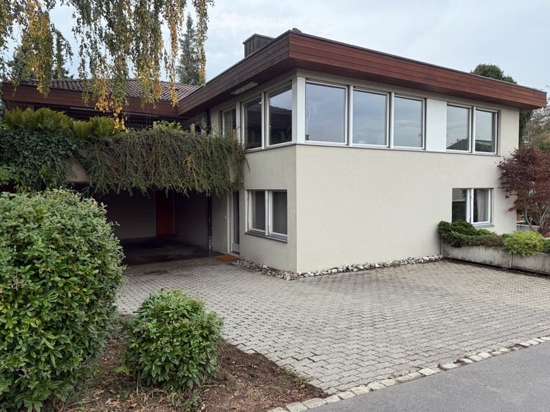 Modern two-story house with a flat roof, large windows, and a paved driveway. The house features a garage entrance and is surrounded by well-maintained shrubs and trees.