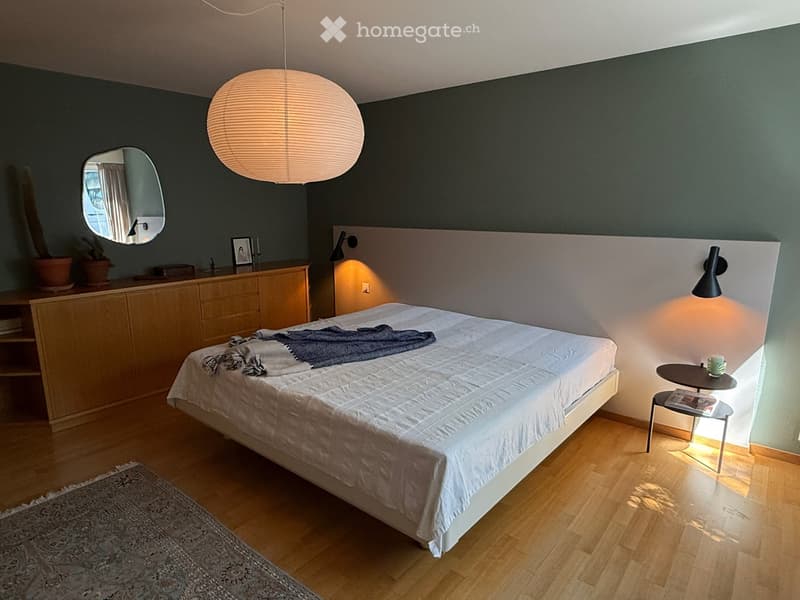 Modern bedroom with a large bed, white bedding, a nightstand with a lamp, a round pendant light, a mirror, and a small side table with a cup.
