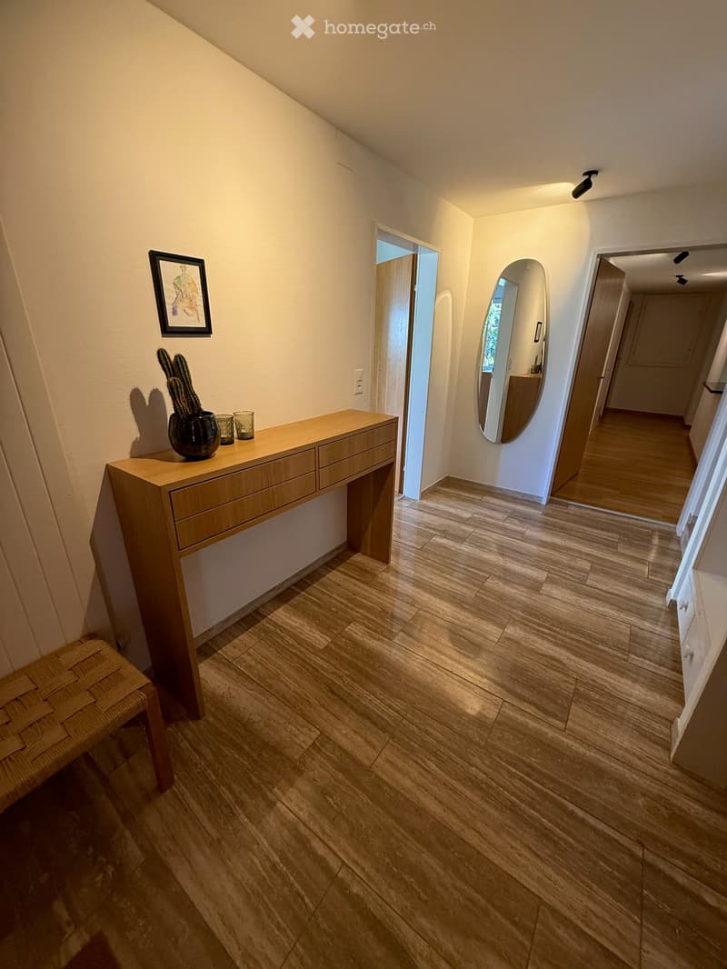 A modern hallway with wooden flooring, a wooden console table with a vase and glasses, a framed picture, and a mirror in an adjacent room.