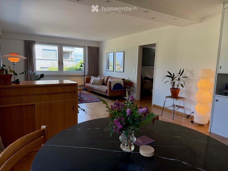 Bright living room with wooden flooring, large windows, a sofa, a round table with a flower arrangement, a tall lamp, and a cabinet.