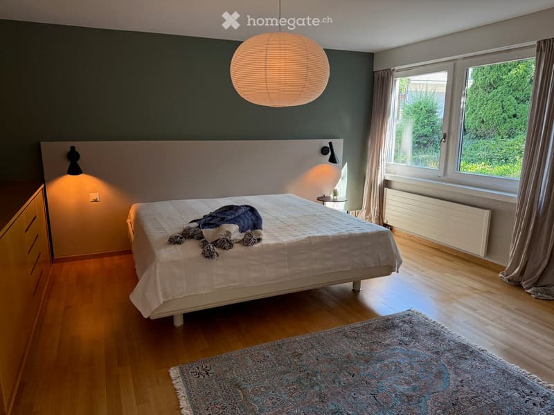 Modern bedroom with wooden flooring, large bed, nightstand, and large window with curtains.