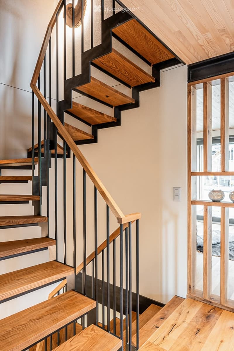Modern wooden staircase with black metal railing, leading to an open area with wooden flooring and a small decorative table in the background.