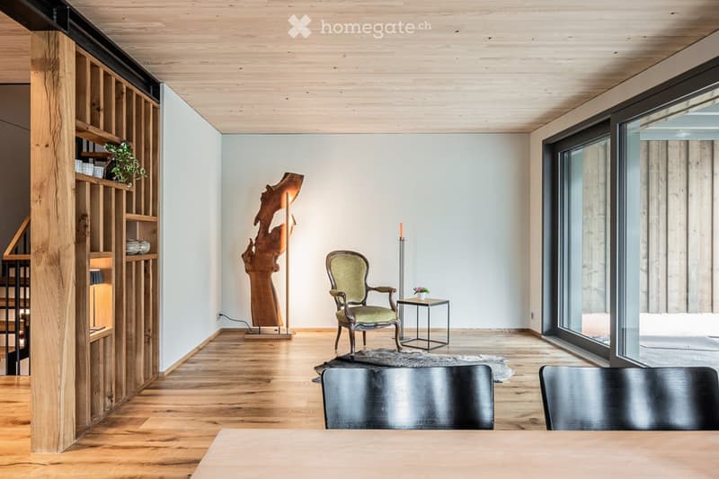 Modern living room with wooden flooring, large windows, a green armchair, a small table, a wooden shelf with decorative items, a floor lamp, and a sculptural piece.