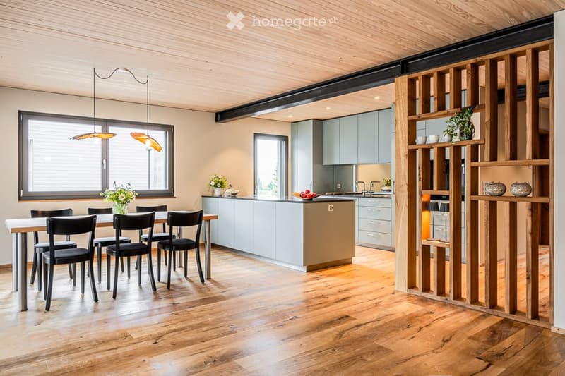Open-plan kitchen and dining area with light blue cabinets, a central island, a dining table with six chairs, pendant lights, and wooden shelves with decorative items.