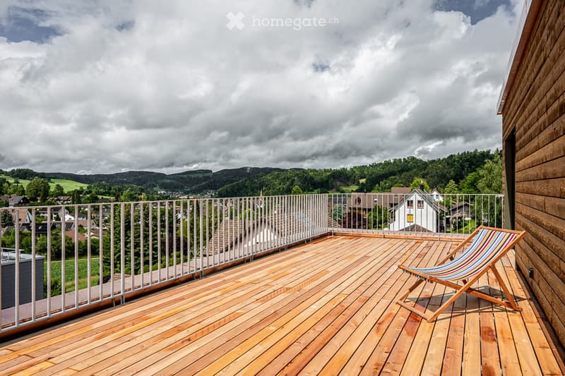 Wooden deck with a lounge chair, overlooking a scenic valley with houses and trees.