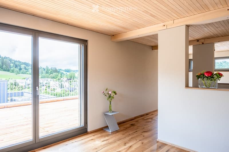 Bright, empty room with wooden flooring, large sliding glass door leading to a balcony, and a view of greenery outside. Minimal decor includes a small table with a vase of flowers.