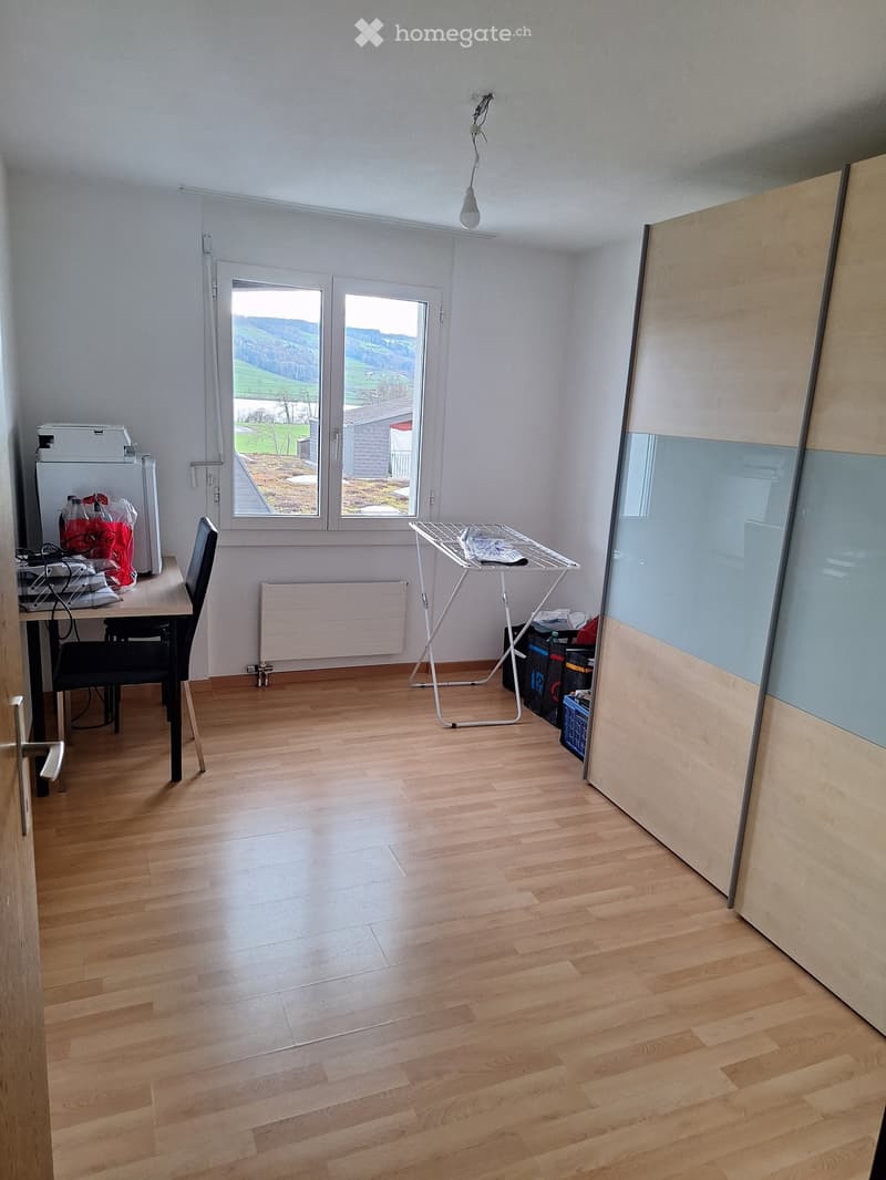 A furnished room with wooden flooring, a large window with a scenic view, a chair, a table with a washing machine, and an ironing board.