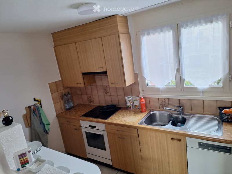 Modern kitchen with wooden cabinets, stainless steel sink, electric stove, and dishwasher. Countertop has various items including bottles, cups, and a frying pan.