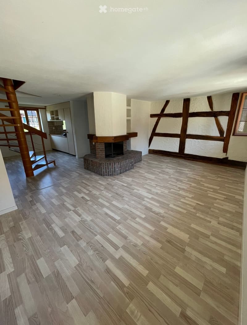 Empty room with wooden flooring, a central fireplace, and a visible staircase leading to another level.