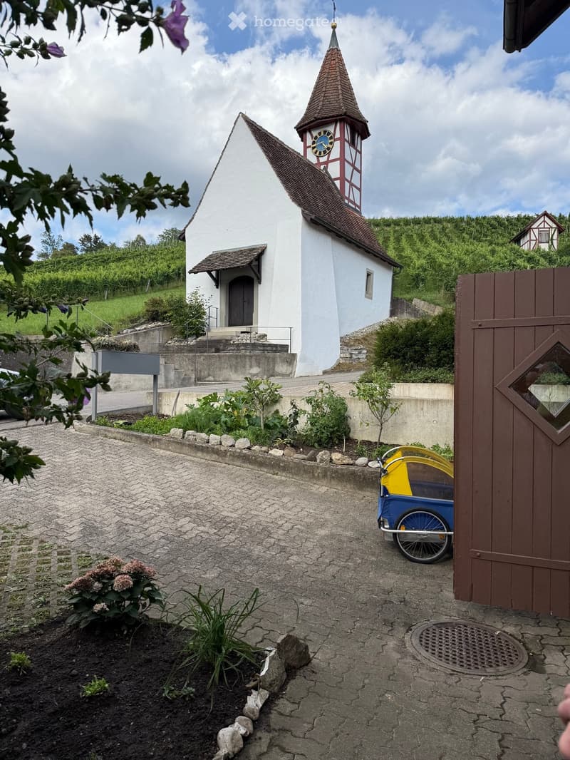 A small white church with a red and white tower and a clock, surrounded by greenery and a paved area with a small blue and yellow cart.