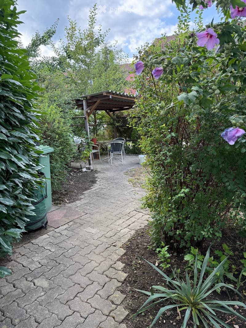 A garden path lined with lush greenery and flowers leads to a small wooden pergola with a table and chairs, surrounded by dense vegetation and a partially visible house in the background.