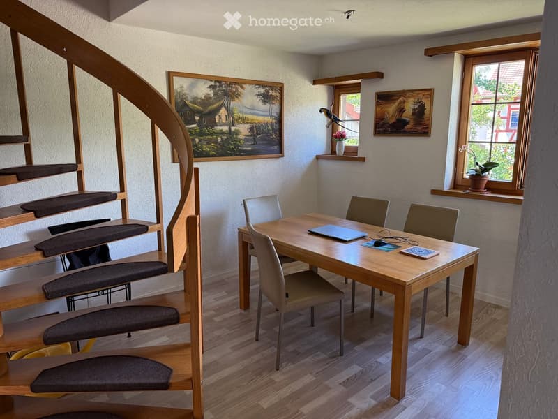 A cozy room with a wooden table, four chairs, a laptop, a mouse, a small stool, and artwork on the walls. The room features wooden flooring and a curved staircase with dark cushions.