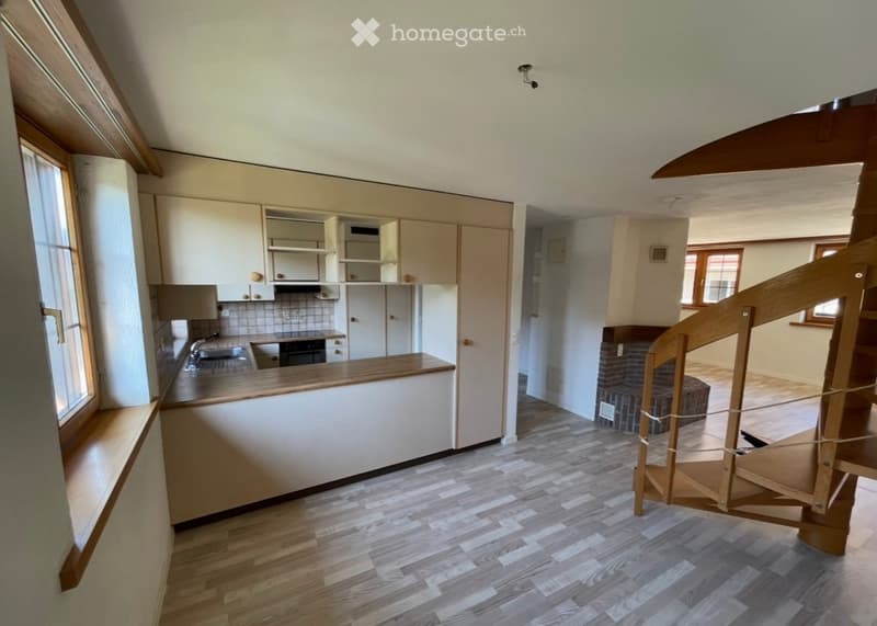 Open-plan kitchen and living area with light wood flooring, white kitchen cabinets, a central island, and a wooden staircase leading to an upper level. A brick fireplace is visible in the living area.