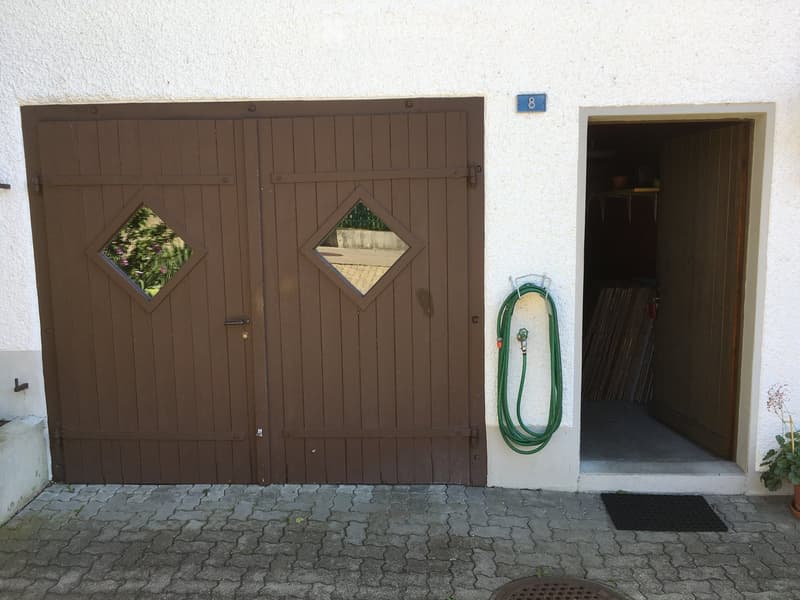 Exterior view of a building with a large wooden garage door featuring diamond-shaped windows, a green garden hose, and a small open doorway leading to a storage area.