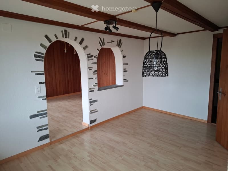 Empty room with wooden flooring, white walls, and a decorative arched doorway. Features a hanging pendant light and exposed wooden ceiling beams.