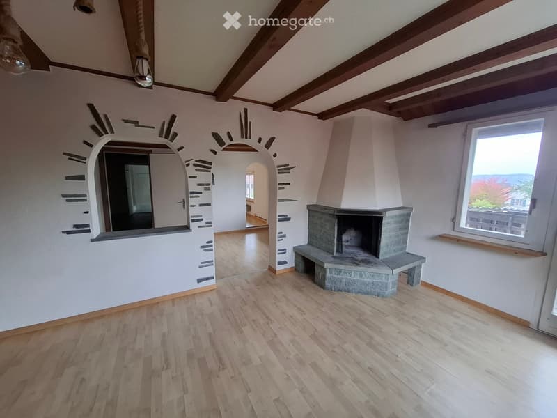 Empty room with wooden flooring, exposed wooden beams on the ceiling, a fireplace with a stone surround, and a large window with a scenic view.