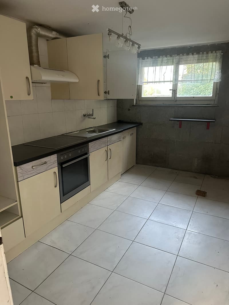 Empty kitchen with white tiled floor, light-colored cabinets, and a black countertop. Features include an electric stove, sink, and a window with white curtains.
