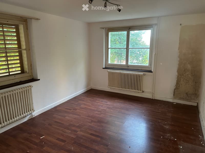 Empty room with wooden flooring, white walls, two windows with radiators below, and a partially painted wall.
