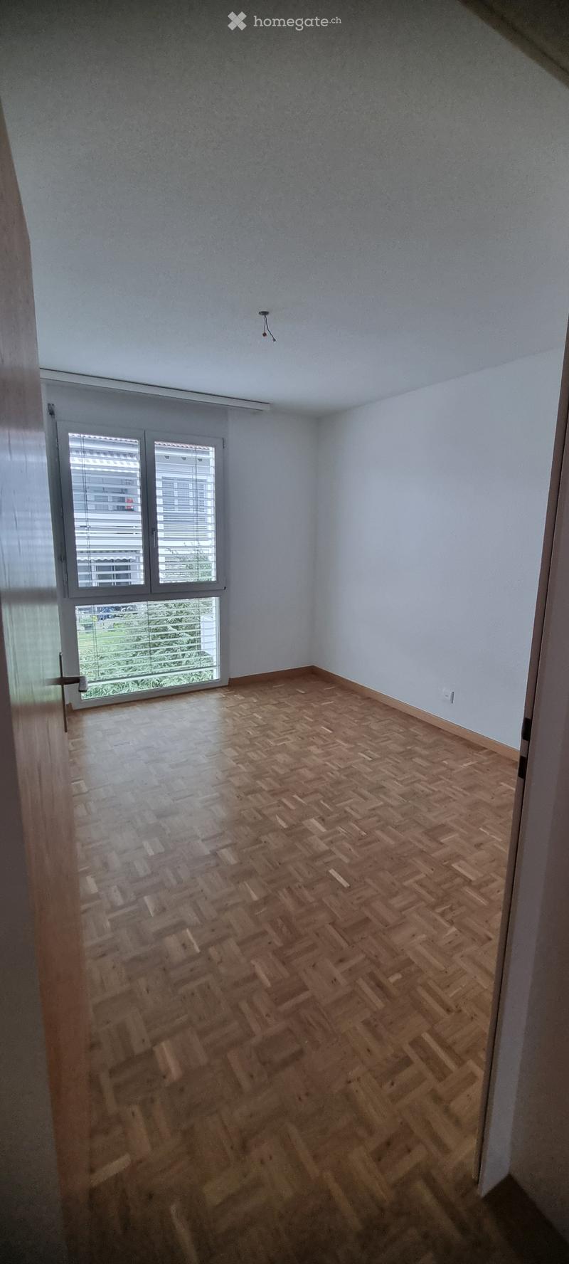 Empty room with wooden herringbone flooring, white walls, and large windows with blinds. No furniture or equipment present.