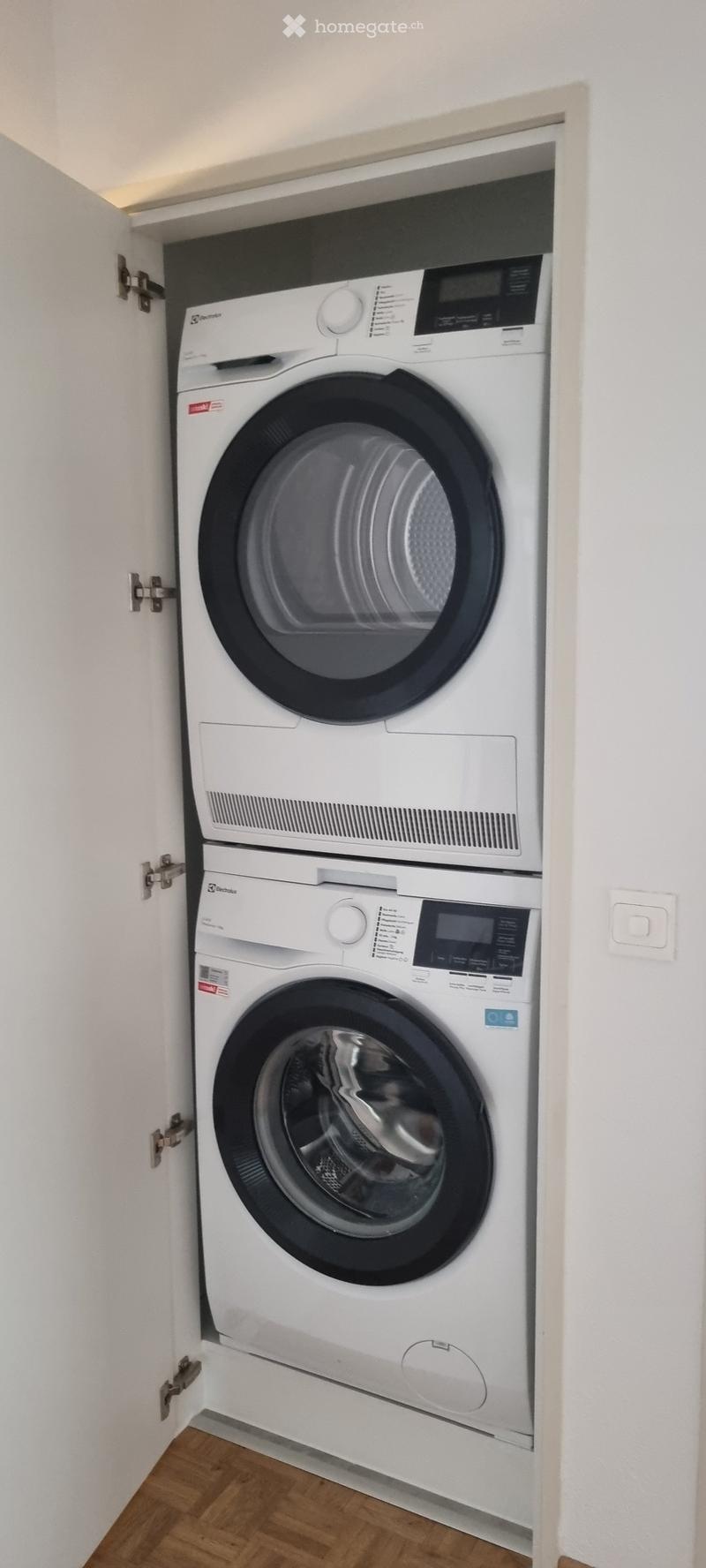 A compact, modern laundry area with a stacked washing machine and dryer unit, white walls, and wooden flooring.