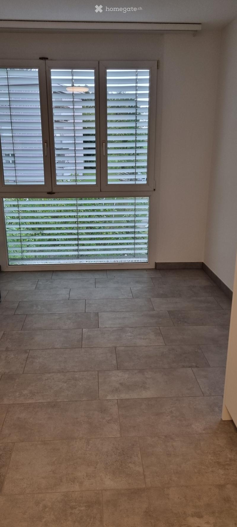 Empty room with tiled floor and large windows with blinds, allowing natural light to enter.