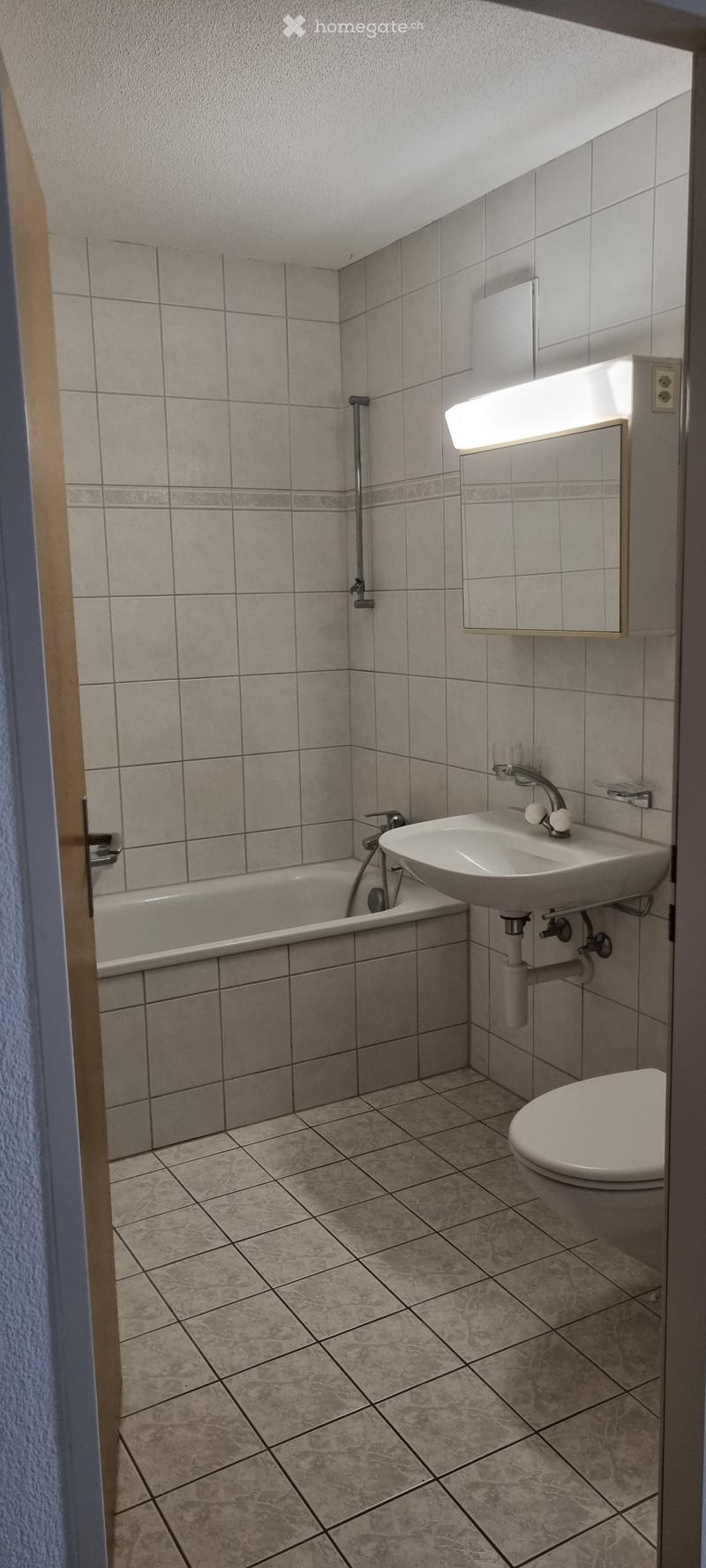 A small bathroom with tiled walls and floor, featuring a bathtub, sink, toilet, and a mirror with a built-in light.