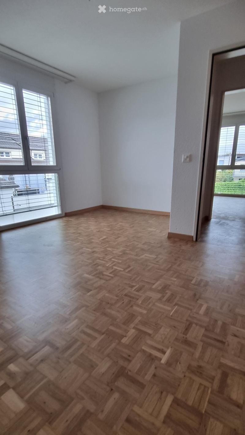 Empty room with wooden herringbone flooring, white walls, large windows with blinds, and a sliding glass door leading to a balcony.