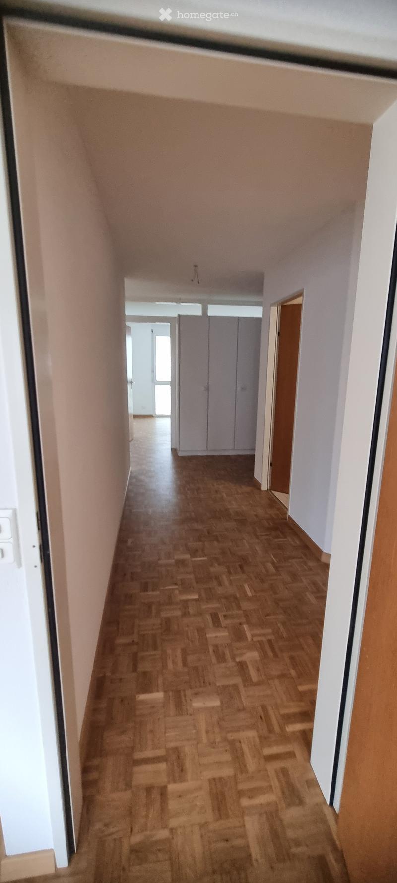 A narrow hallway with wooden flooring and white walls, leading to an open area with a door and closet visible at the end.