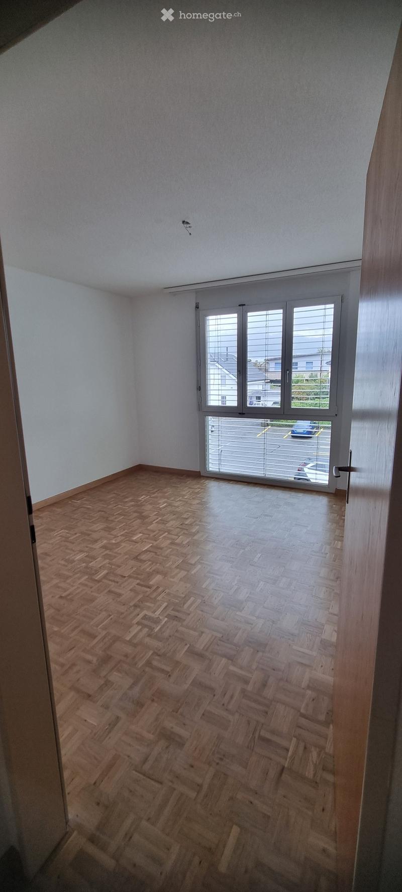 Empty room with wooden herringbone flooring, white walls, and large window with blinds overlooking a parking lot.