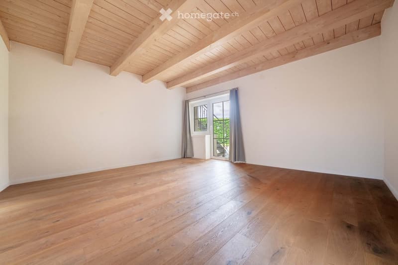 Empty room with wooden flooring and exposed wooden ceiling beams. Features a large window with grey curtains and a sliding glass door leading outside.