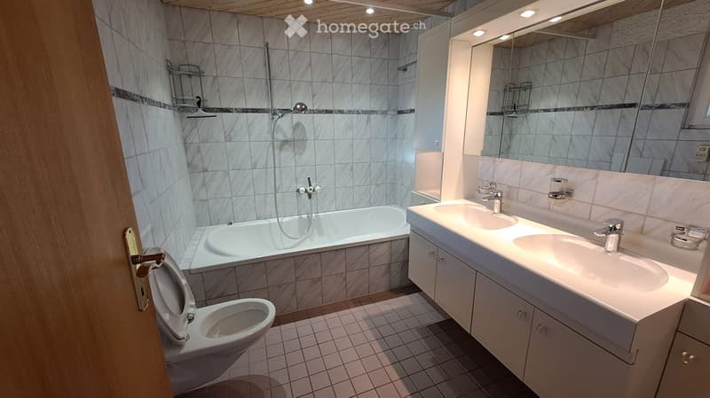 Modern bathroom with marble tiles, a bathtub, a toilet, a double sink vanity, and a large mirror. The shower area is visible with a glass enclosure and a handheld showerhead.