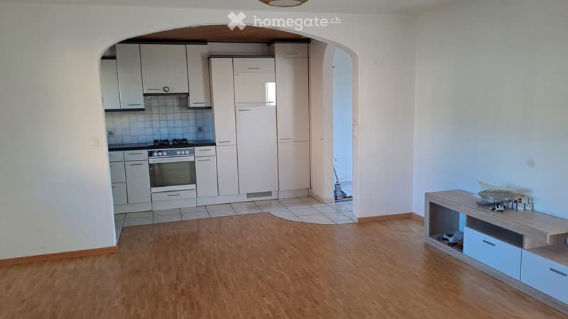 A modern, open-plan living and dining area with a wooden floor, white cabinets, and a built-in oven in the kitchen area. A small table with drawers is positioned near the living space.