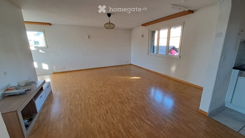 Empty room with wooden flooring, white walls, and large windows allowing natural light. There is a small cabinet with drawers on the left side and a partial view of a kitchen area on the right.
