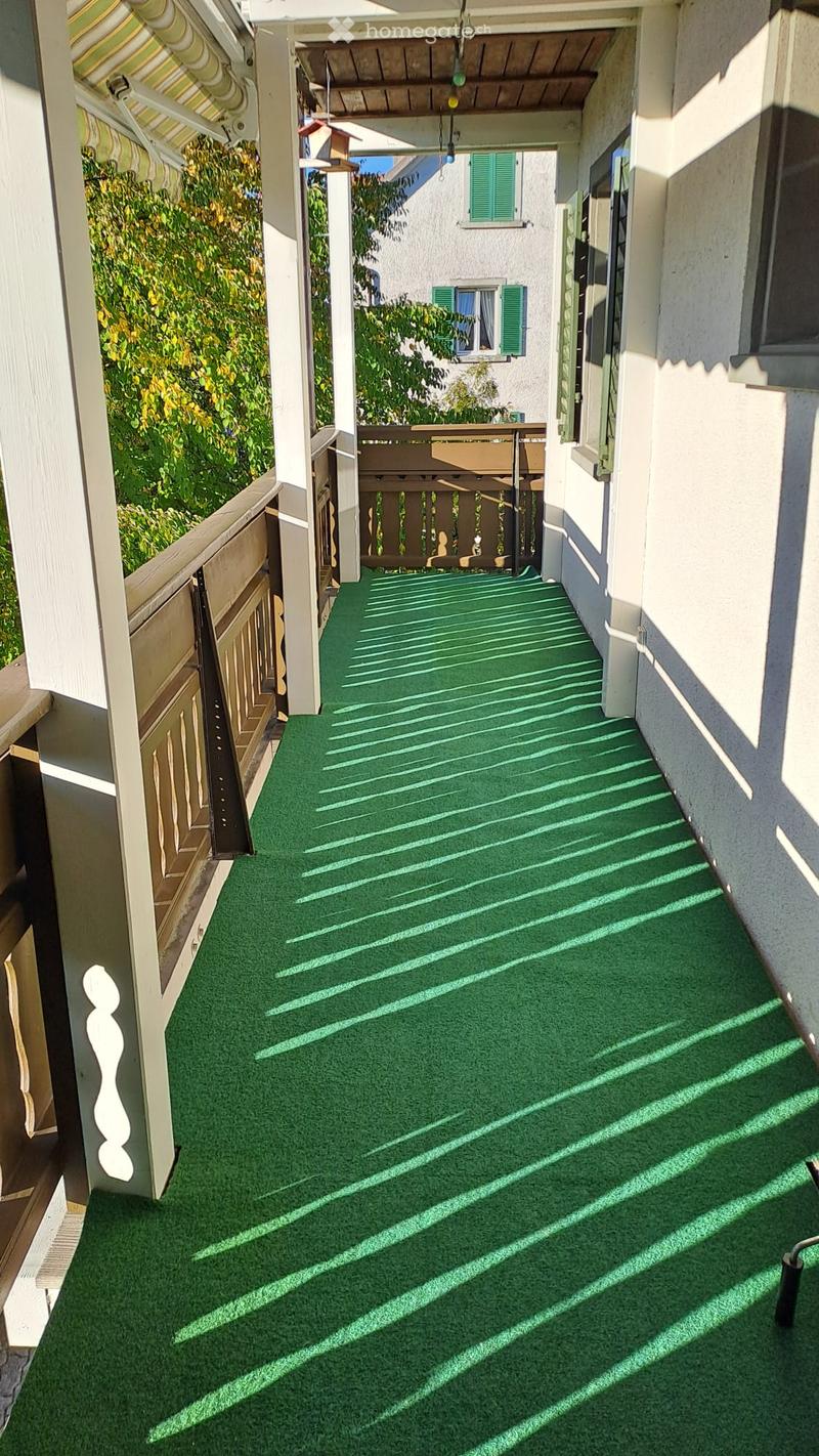 Green artificial turf balcony with wooden railings, white columns, and striped awning.