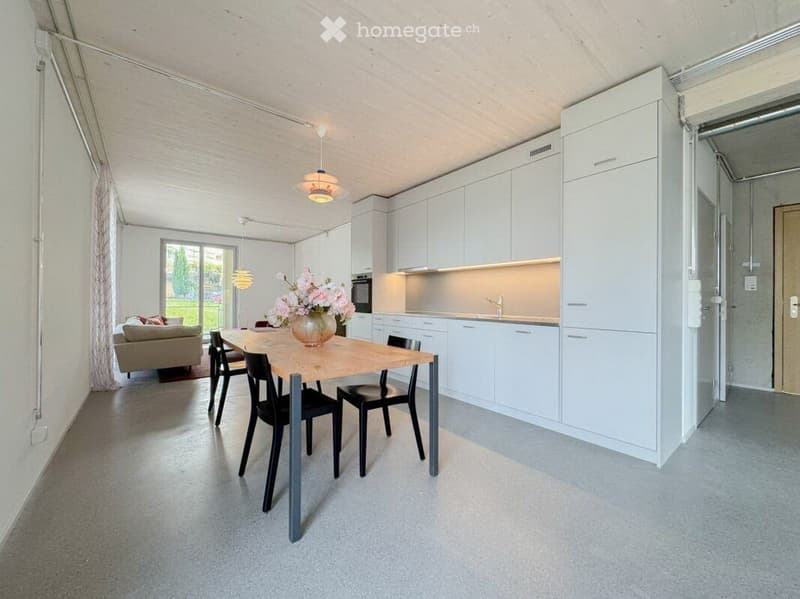 Open-plan living and dining area with a sofa, dining table, chairs, and a modern kitchen with white cabinets and stainless steel appliances.