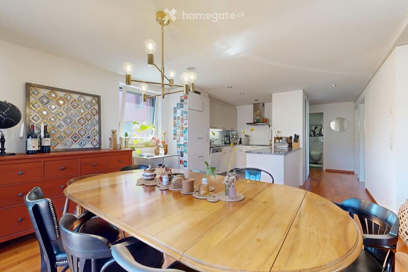 A well-lit dining area with a wooden dining table surrounded by chairs, a cabinet with bottles and decorative items, and a modern kitchen in the background.