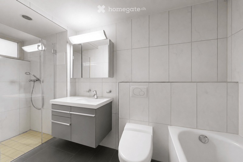 Modern bathroom with white tiles, a sink with a mirror, a toilet, a bathtub, and a glass-enclosed shower area.